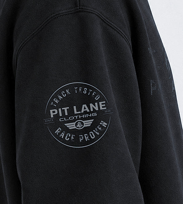 Close-up of a black sweatshirt sleeve featuring a circular gray graphic print on the upper arm that reads 'PIT LANE CLOTHING' with 'TRACK TESTED' and 'RACE PROVEN'.