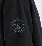 Close-up of a black sweatshirt sleeve featuring a circular gray graphic print on the upper arm that reads 'PIT LANE CLOTHING' with 'TRACK TESTED' and 'RACE PROVEN'.
