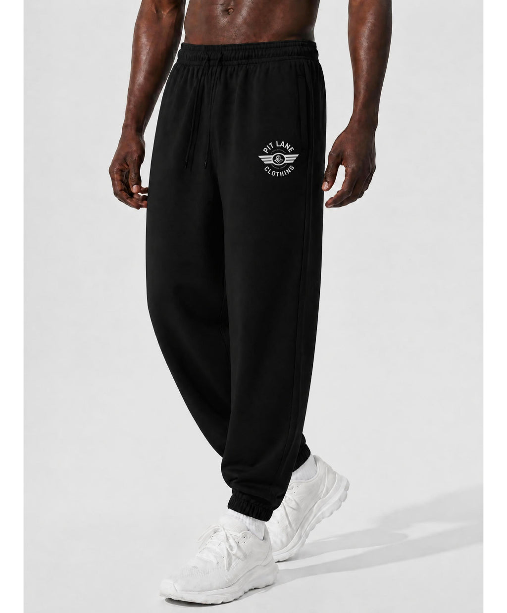 A man wearing black jogger sweatpants with a white 'PIT LANE CLOTHING' graphic on the left thigh, paired with white sneakers, shown from the waist down against a light gray studio background.