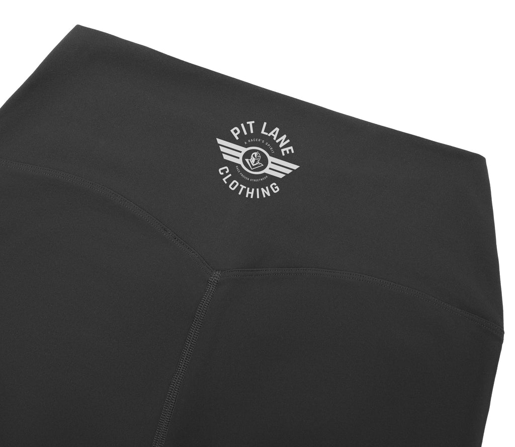 Close-up of black athletic leggings with a white circular winged logo reading 'PIT LANE CLOTHING' on the upper left hip, shown against a white background.