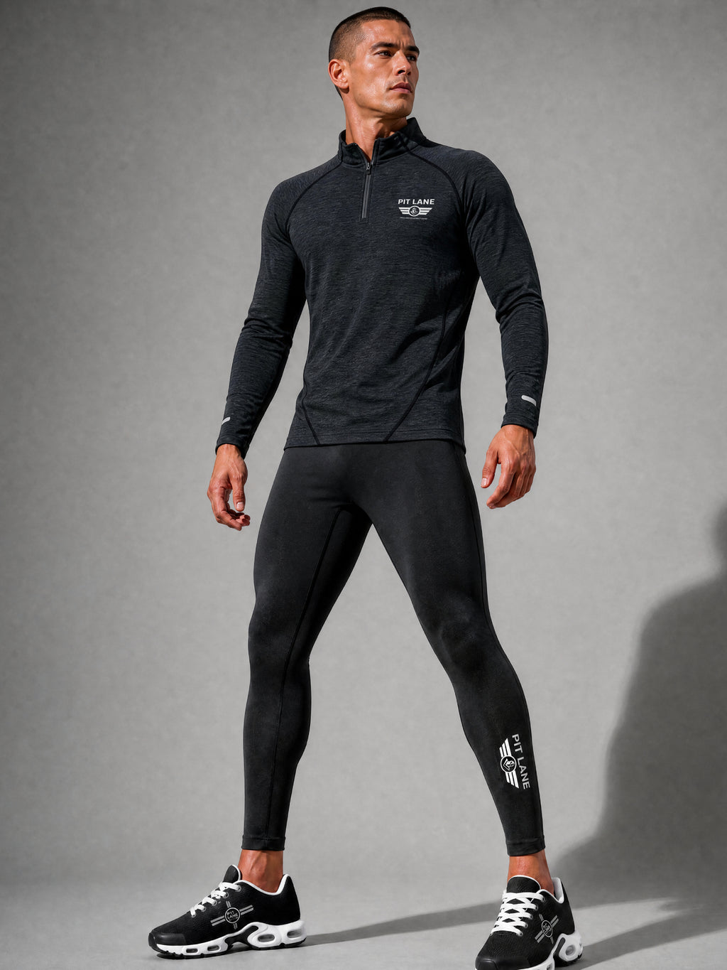 A man modeling a dark gray long-sleeve quarter-zip athletic top and matching black leggings with white PIT LANE CLOTHING logo details, standing against a gray studio background.