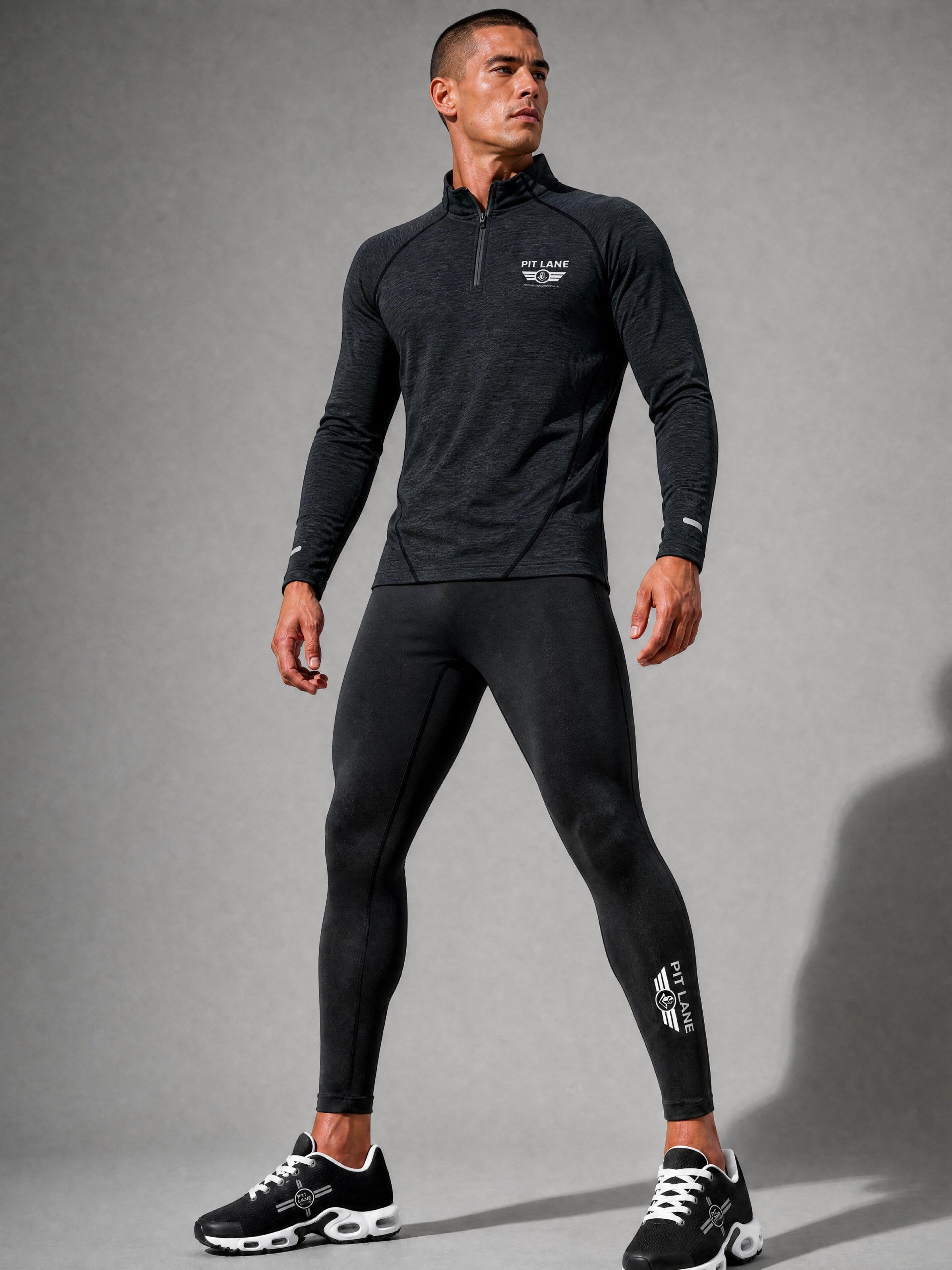 A man modeling a dark gray long-sleeve quarter-zip athletic top and matching black leggings with white PIT LANE CLOTHING logo details, standing against a gray studio background.