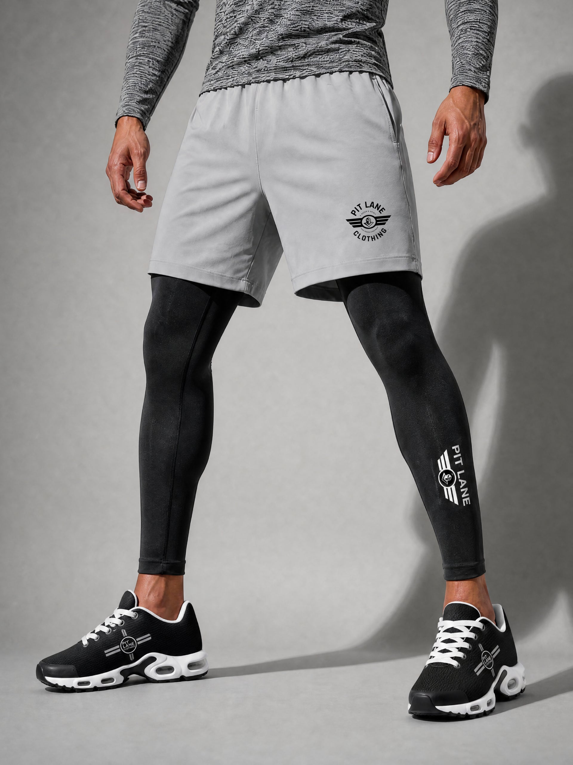 A model wearing light gray athletic shorts with a black PIT LANE CLOTHING logo, layered over black compression leggings and black sneakers, shown from the waist down against a gray studio background.