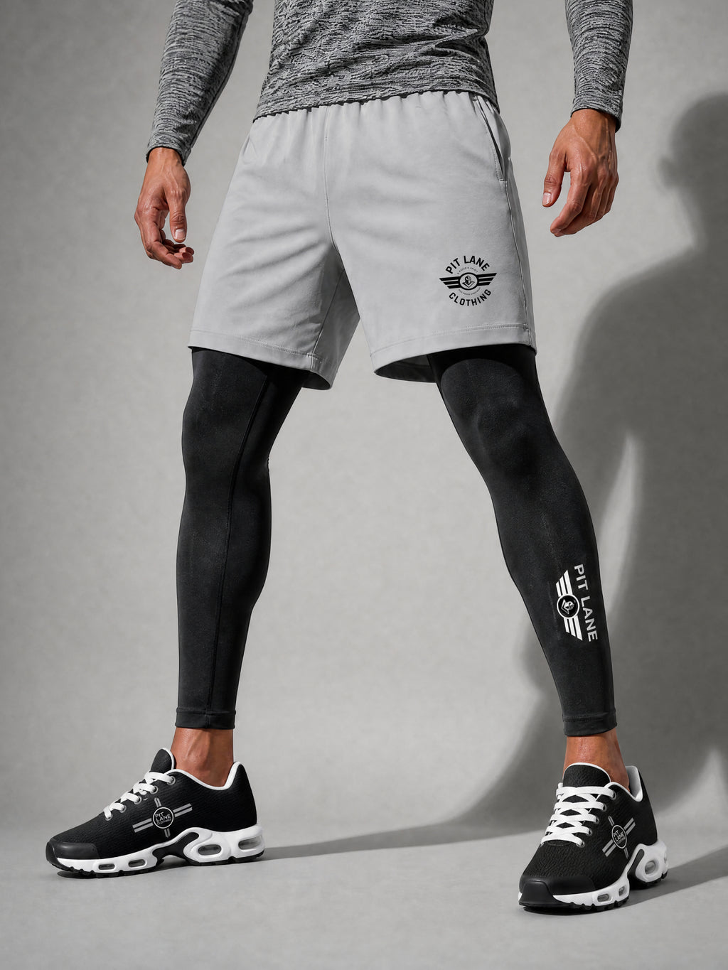 A model wearing light gray athletic shorts with a black PIT LANE CLOTHING logo, layered over black compression leggings and black sneakers, shown from the waist down against a gray studio background.