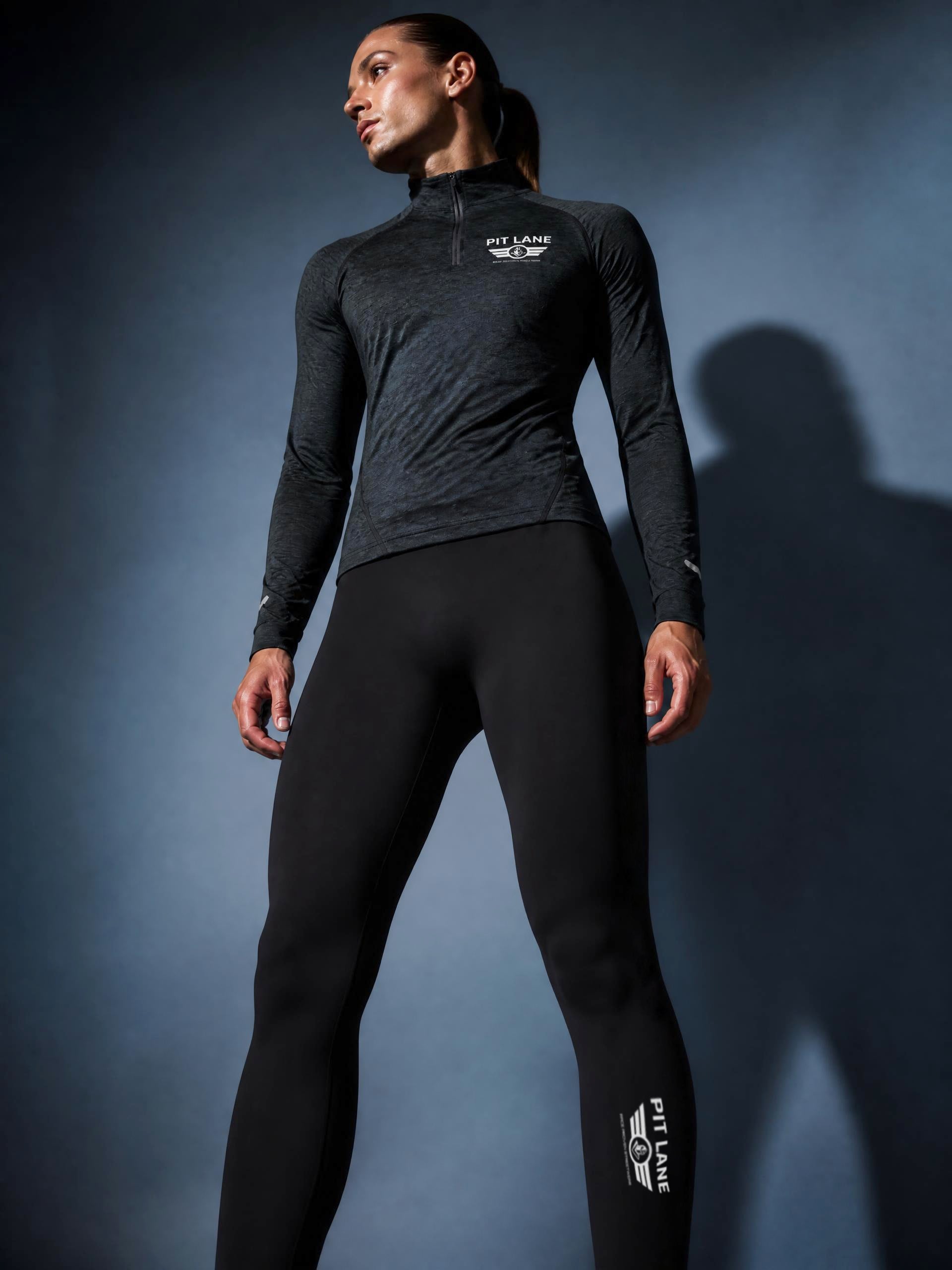 A woman modeling a dark gray heathered long-sleeve quarter-zip athletic top and black leggings with white PIT LANE CLOTHING branding, standing against a gray studio background.
