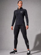 A woman modeling a dark gray heathered long-sleeve quarter-zip athletic top and black leggings with white PIT LANE CLOTHING branding, standing against a gray studio background.