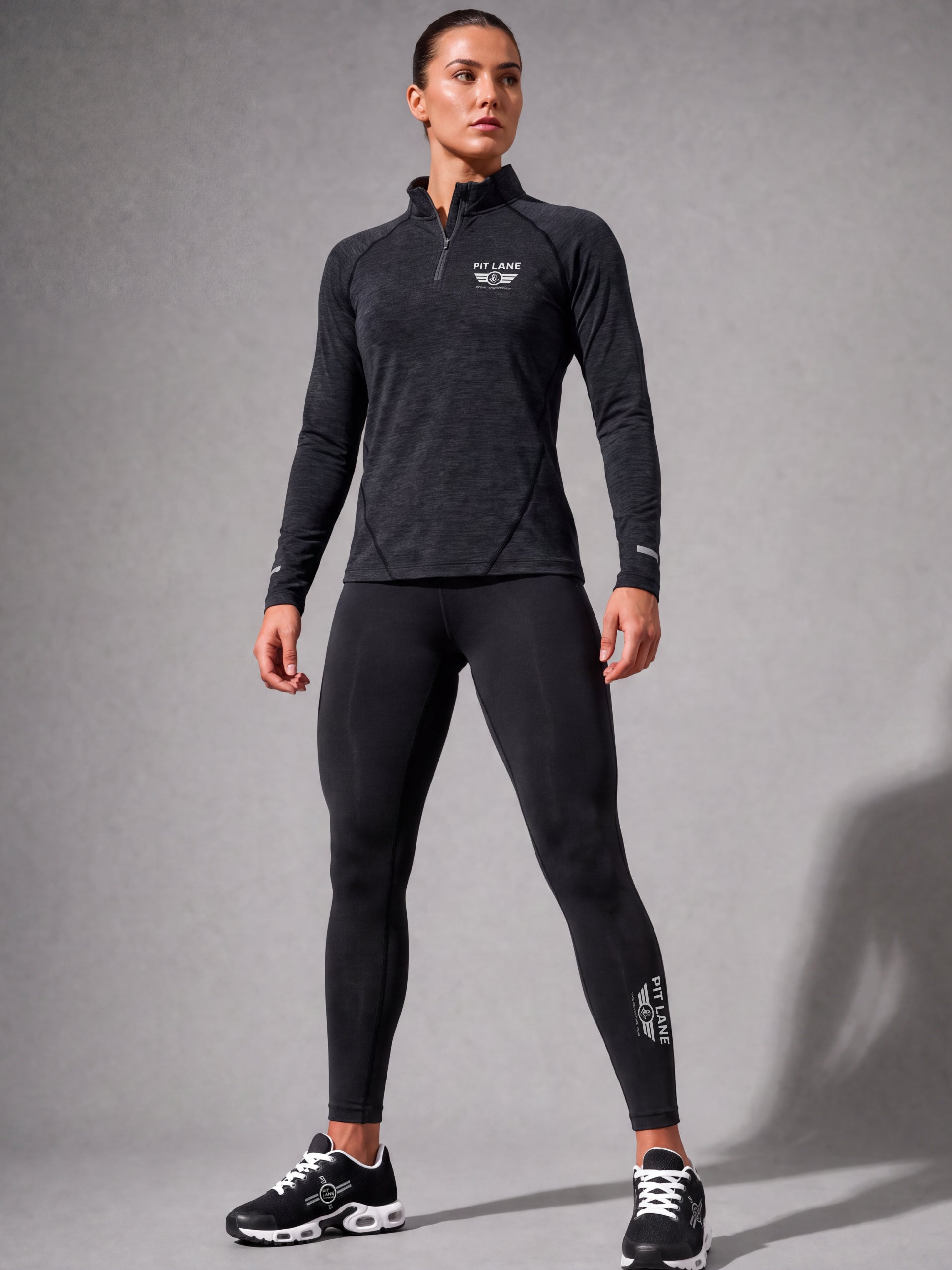 A woman modeling a dark gray heathered long-sleeve quarter-zip athletic top and black leggings with white PIT LANE CLOTHING branding, standing against a gray studio background.
