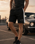 A man wearing black athletic shorts with a white side stripe and a white 'PIT LAKE CLOTHING' logo on the left leg, standing next to a classic car on an outdoor road during sunset.