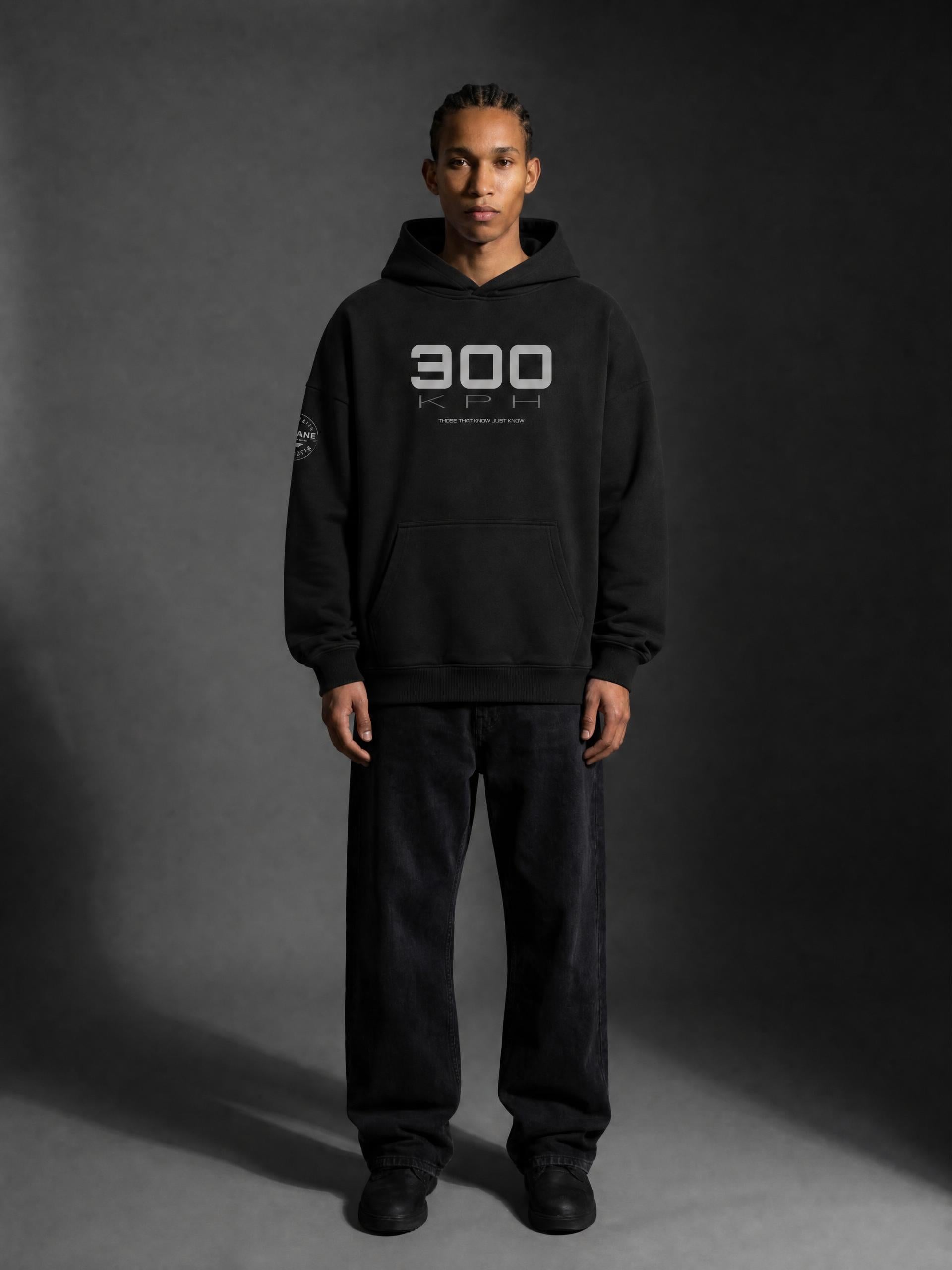A man wearing a PIT LANE CLOTHING black pullover hoodie with a white '300 KPH' graphic on the chest, paired with black sweatpants and black sneakers, standing against a dark gray studio background.