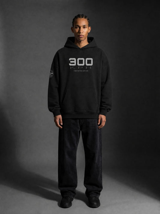 A man wearing a PIT LANE CLOTHING black pullover hoodie with a white '300 KPH' graphic on the chest, paired with black sweatpants and black sneakers, standing against a dark gray studio background.