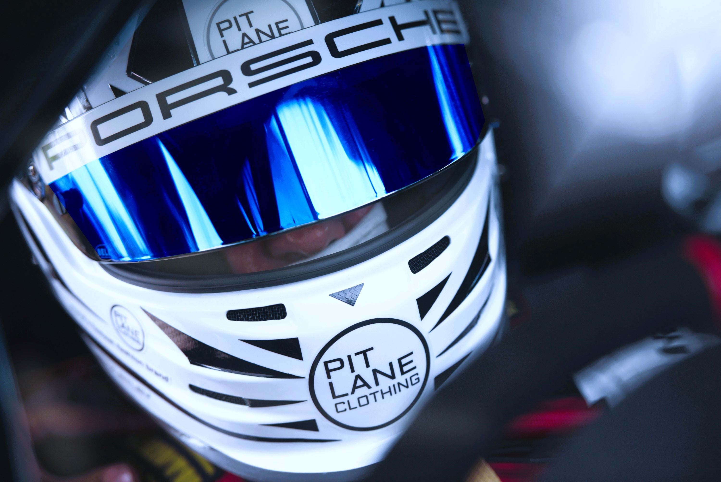 Close-up of a white racing helmet with a reflective blue visor, featuring visible 'Porsche' branding and a 'Pit Lane Clothing' logo on the lower front.