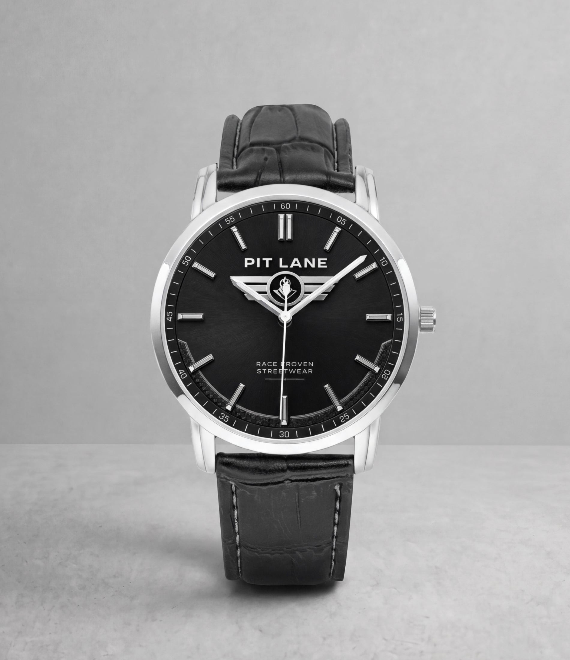 A Pit Lane wristwatch with a black dial, silver-tone case, and black leather strap, centered against a neutral gray background.