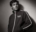 A man wearing a black track jacket with white side stripes and a white 'PIT LANE CLOTHING' logo on the chest, photographed in a dramatic studio portrait against a dark gray background.