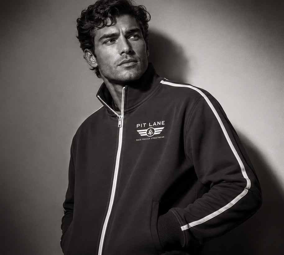 A man wearing a black track jacket with white side stripes and a white 'PIT LANE CLOTHING' logo on the chest, photographed in a dramatic studio portrait against a dark gray background.