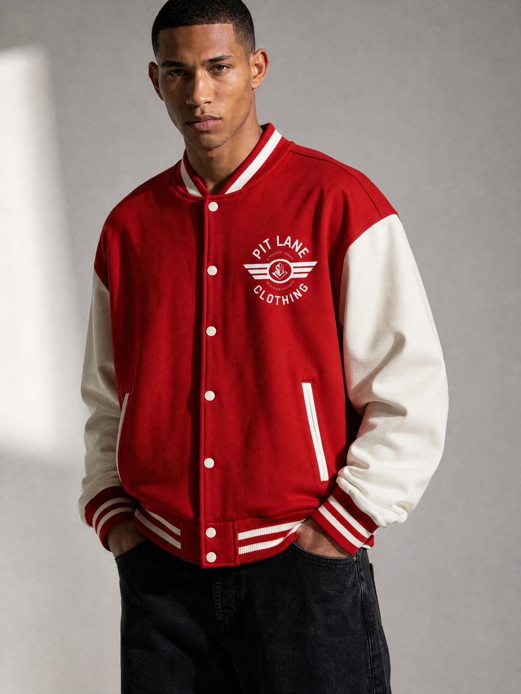 A man wearing a red and off-white varsity jacket with 'PIT LAKE CLOTHING' embroidered on the chest, posed against a gray studio background.