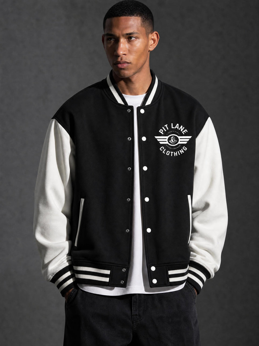 A man wearing a black and white varsity jacket with 'PIT LANE CLOTHING' embroidered on the chest, posed against a dark gray studio background.