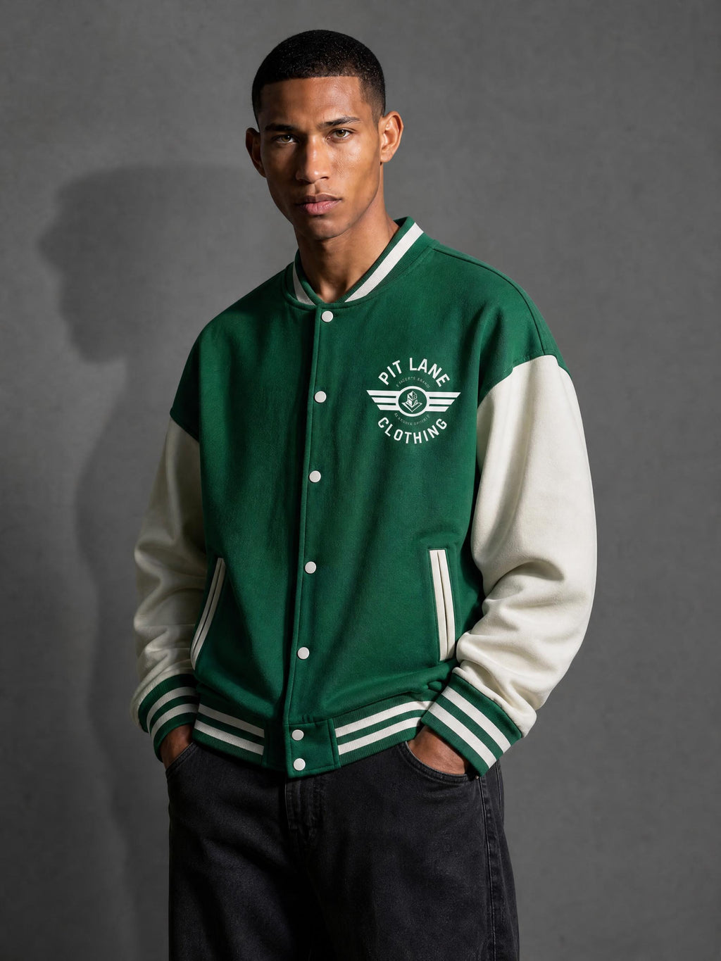 A man wearing a green and cream varsity jacket with 'PIT LANE CLOTHING' printed on the chest, posed against a dark gray studio background.