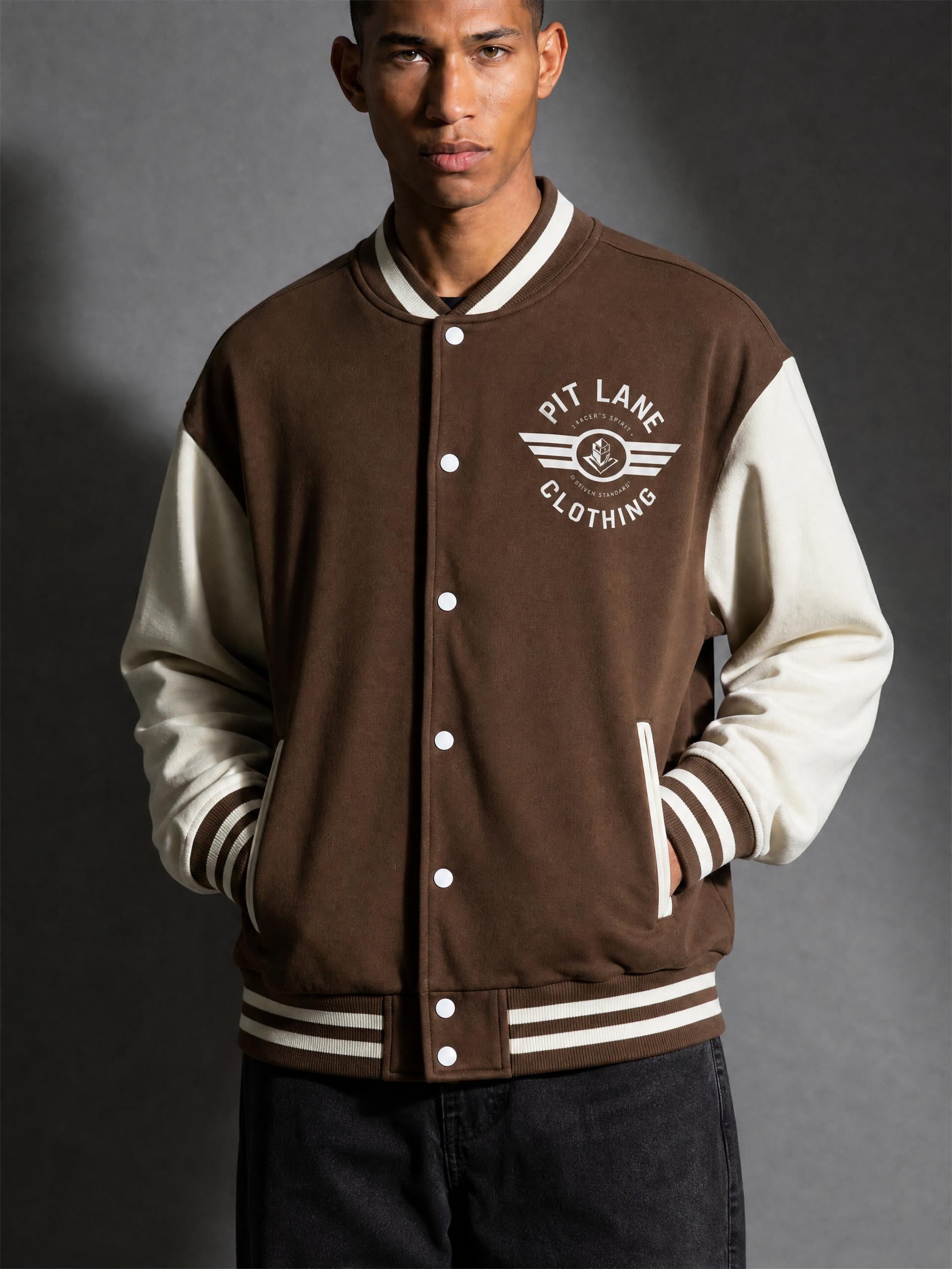 A man wearing a brown and cream varsity jacket with 'PIT LANE CLOTHING' printed on the chest, posed against a dark gray studio background.