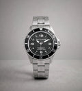 A Pit Lane Clothing stainless steel wristwatch with a black dial and bezel, featuring silver-tone metal case and link bracelet, photographed against a neutral gray background.