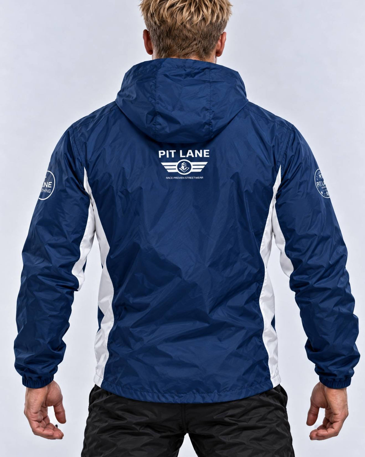 A man viewed from the back wearing a navy blue hooded windbreaker jacket with white side panels and a white 'PIT LANE' logo printed on the upper back.
