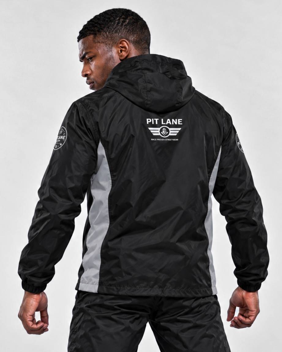 A man wearing a black hooded windbreaker jacket with gray side panels and white 'PIT LANE' branding on the back, shown from behind against a light gray studio background.