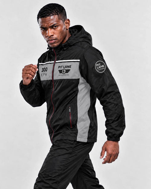 A man wearing a black and gray hooded windbreaker jacket with 'PIT LANE' branding on the chest and sleeve, paired with black pants, posed against a light gray studio background.