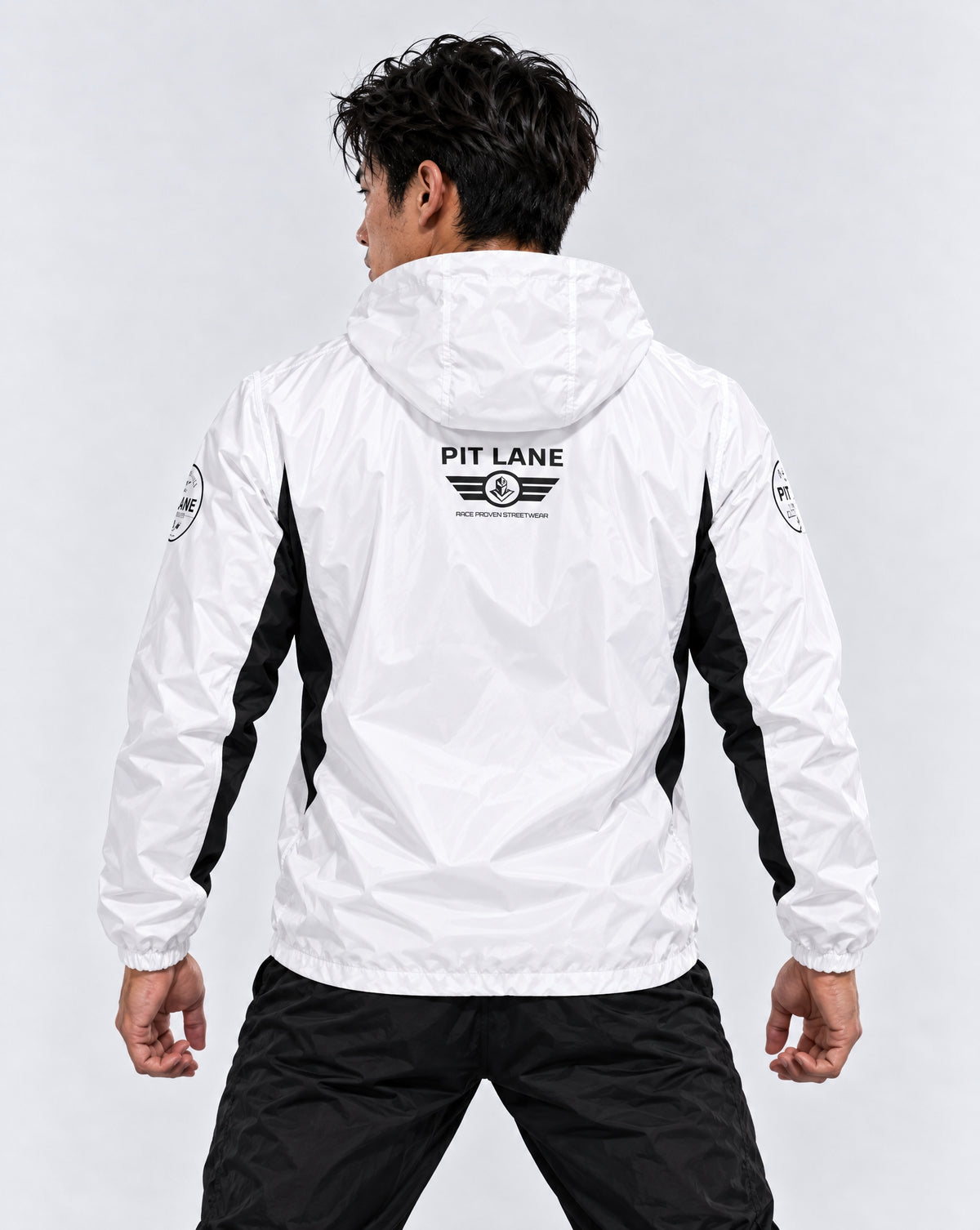 A man viewed from behind wearing a white hooded windbreaker jacket with black side panels and a black 'PIT LANE CLOTHING' logo printed on the back, paired with black pants against a light gray studio background.