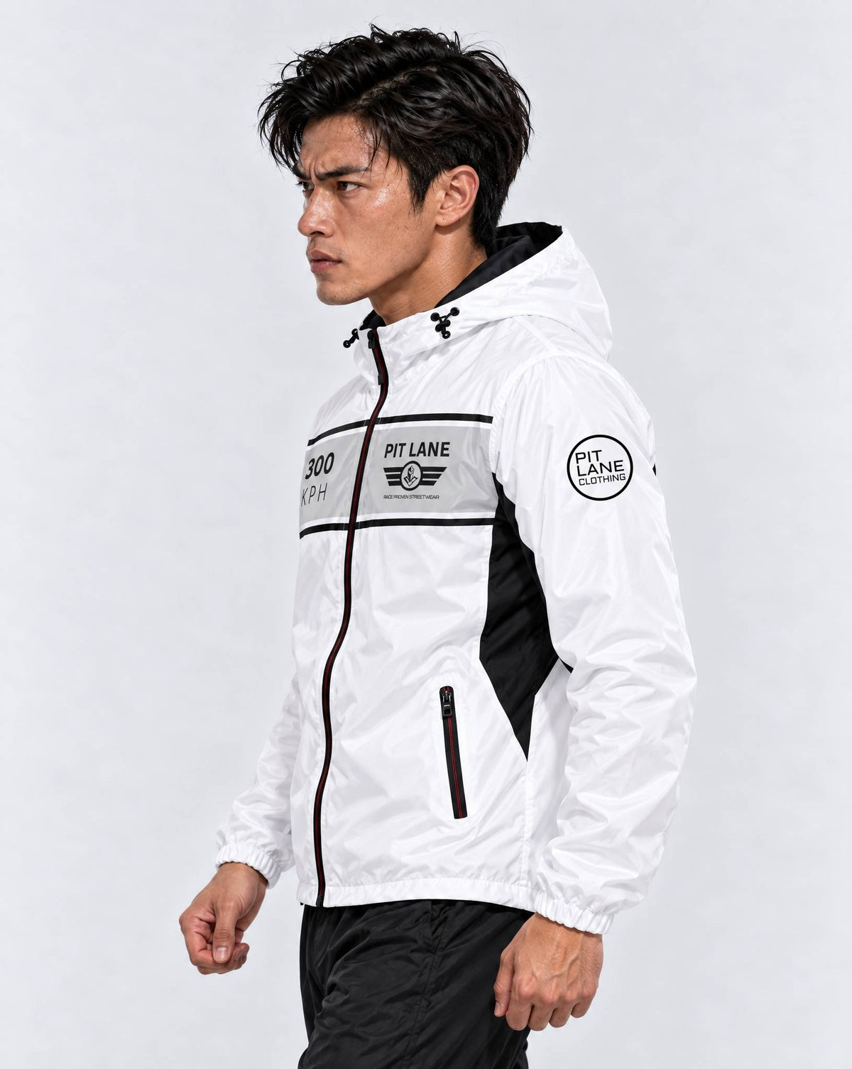 A man wearing a white hooded windbreaker jacket with black and gray racing-style graphics and 'PIT LANE CLOTHING' branding, paired with black pants, against a light gray studio background.