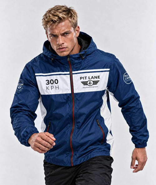 A man wearing a navy blue and white hooded windbreaker jacket with 'PIT LANE' and '300 KPH' printed on the chest, posed against a light gray studio background.