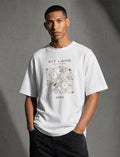 A man wearing a white short-sleeve graphic t-shirt with a brown geometric design and text reading 'PIT LANE CLOTHING' and 'APEX', paired with dark jeans, standing against a textured gray studio background.