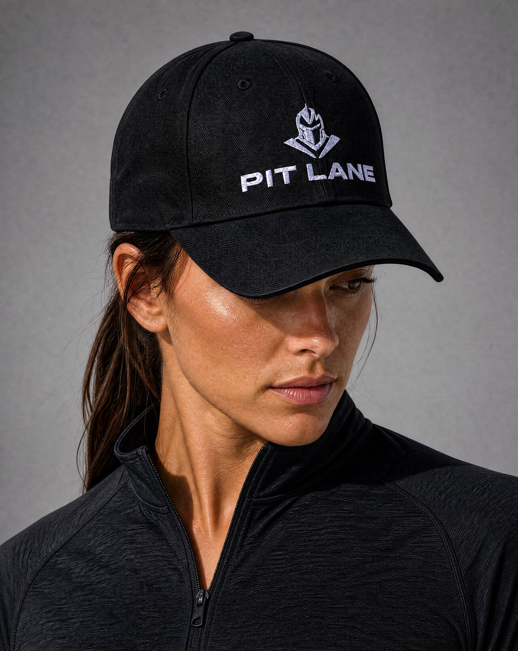 A woman wearing a black baseball cap with a white embroidered PIT LANE logo, paired with a black athletic zip-up jacket, set against a neutral gray background.