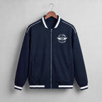 A navy blue zip-up jacket with white contrast stitching, ribbed collar, cuffs, and hem, hanging on a wooden hanger against a light gray textured wall. The left chest features a white circular logo reading 'KIT LANE CLOTHING' with a winged emblem.