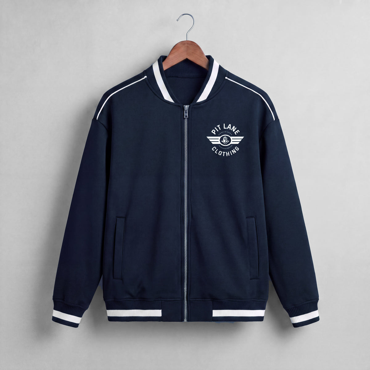 A navy blue zip-up jacket with white contrast stitching, ribbed collar, cuffs, and hem, hanging on a wooden hanger against a light gray textured wall. The left chest features a white circular logo reading 'KIT LANE CLOTHING' with a winged emblem.