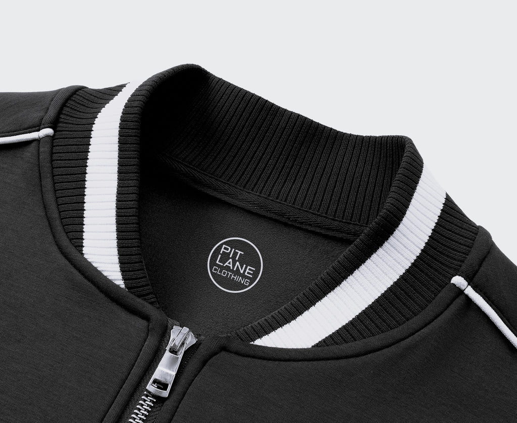 A navy blue zip-up jacket with white contrast stitching, ribbed collar, cuffs, and hem, hanging on a wooden hanger against a light gray textured wall. The left chest features a white circular logo reading 'KIT LANE CLOTHING' with a winged emblem.