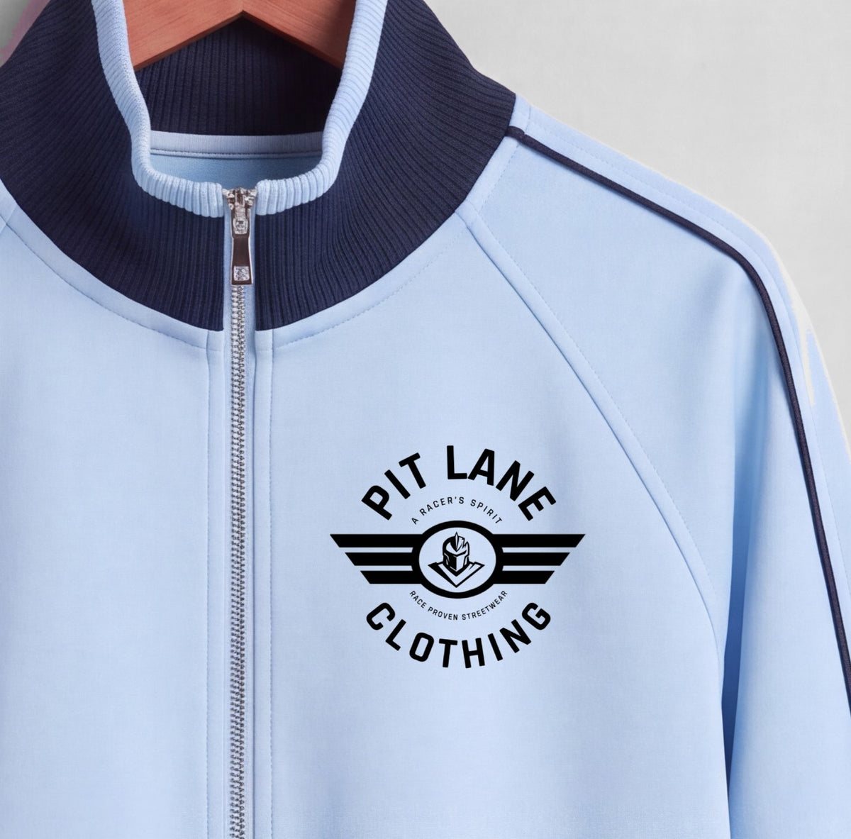 A close-up of a light blue zip-up jacket with a dark navy ribbed collar and a black 'PIT LANE CLOTHING' logo printed on the chest.