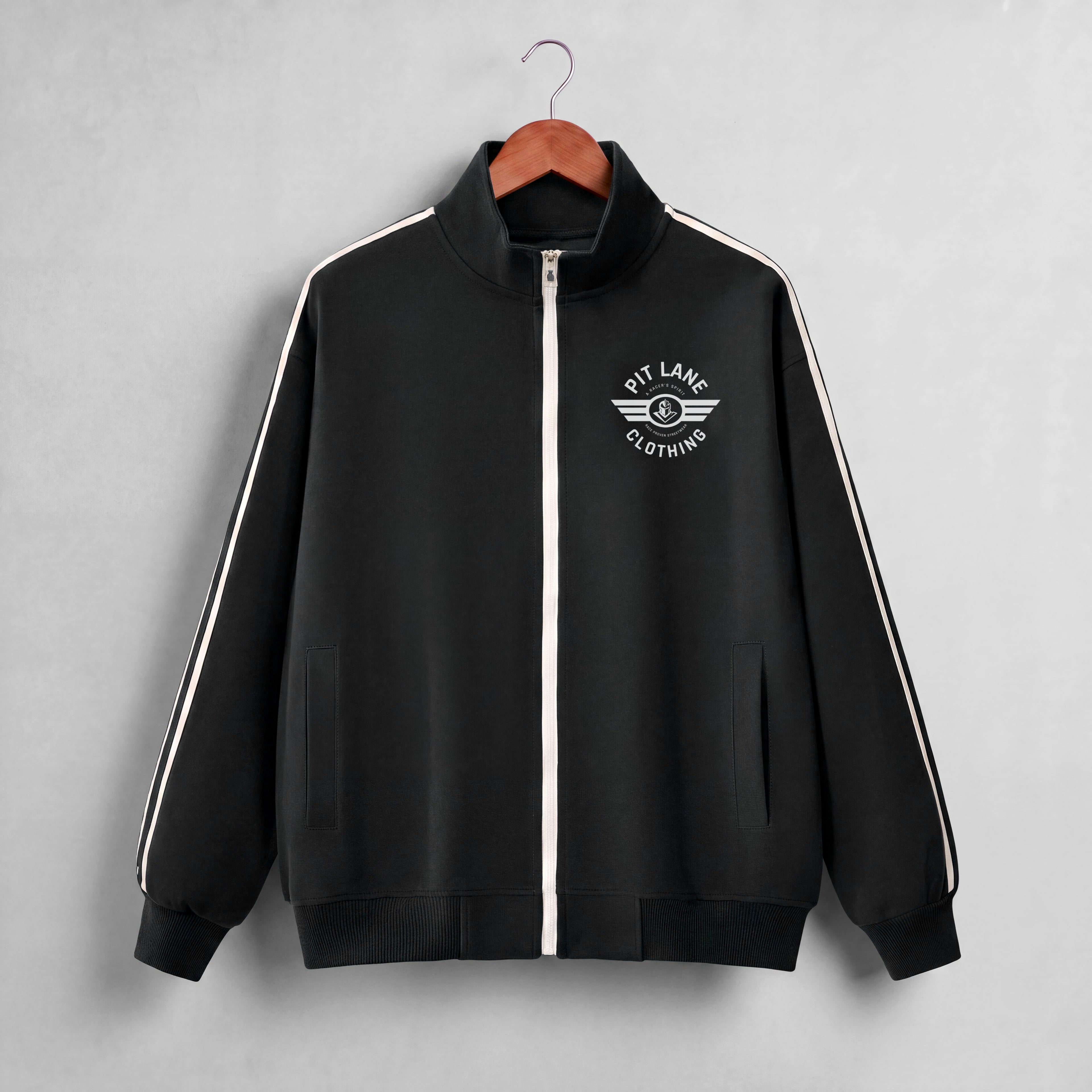 A black zip-up track jacket with a white front zipper, white shoulder stripes, and a white 'PIT LANE CLOTHING' wing logo on the chest, hanging on a wooden hanger against a light gray background.