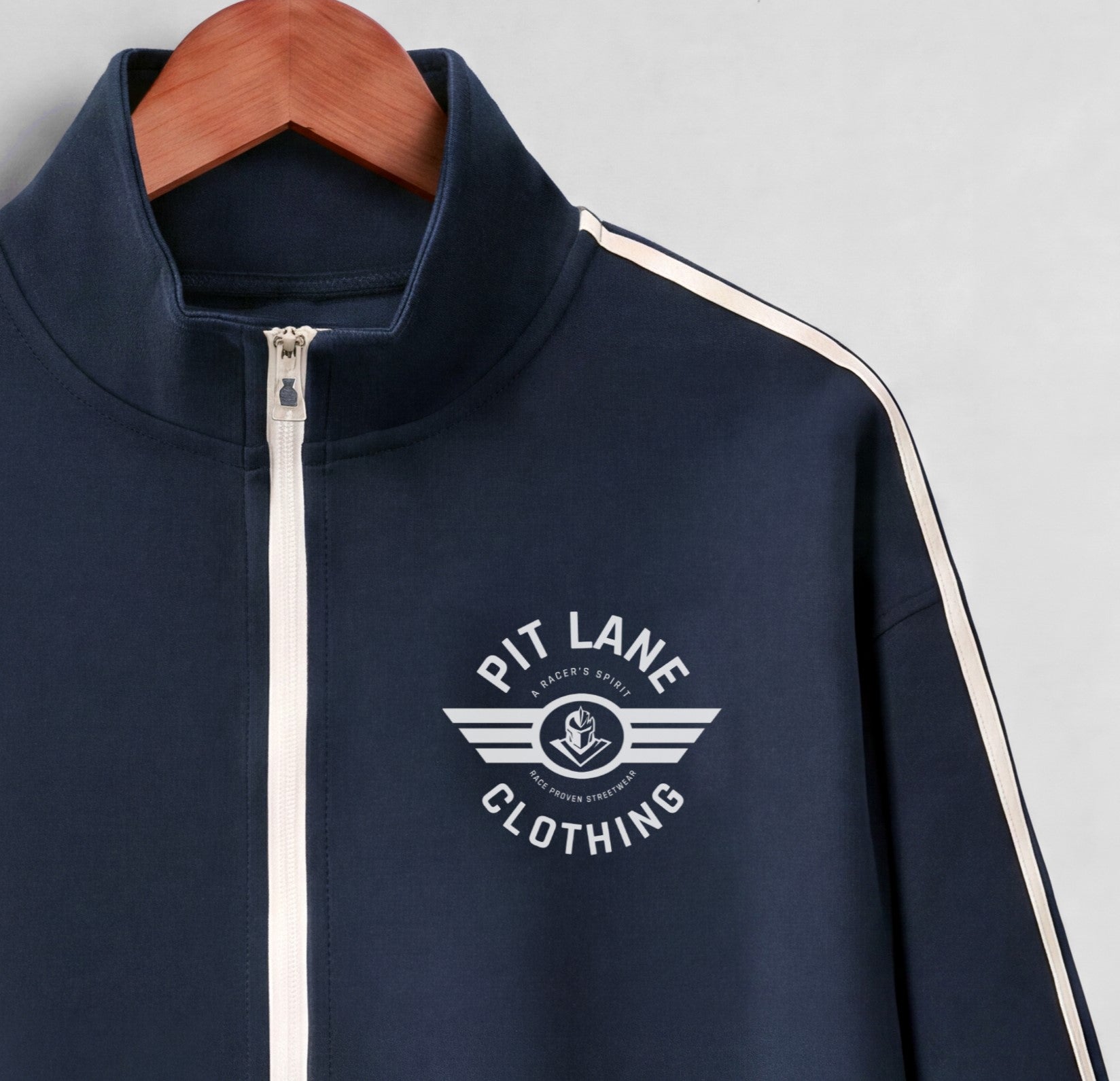 A navy blue zip-up track jacket with white shoulder stripes and a white chest logo reading 'PIT LANE CLOTHING', hanging on a wooden hanger against a light gray background.