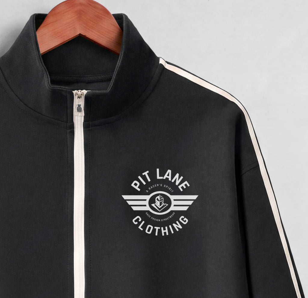 A black zip-up track jacket with a white front zipper, white shoulder stripes, and a white 'PIT LANE CLOTHING' wing logo on the chest, hanging on a wooden hanger against a light gray background.