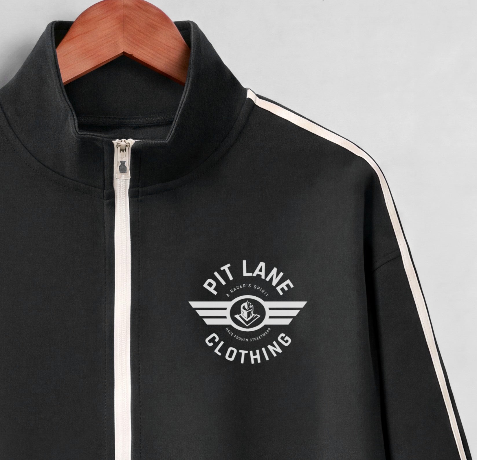 A black zip-up track jacket with a white front zipper, white shoulder stripes, and a white 'PIT LANE CLOTHING' wing logo on the chest, hanging on a wooden hanger against a light gray background.