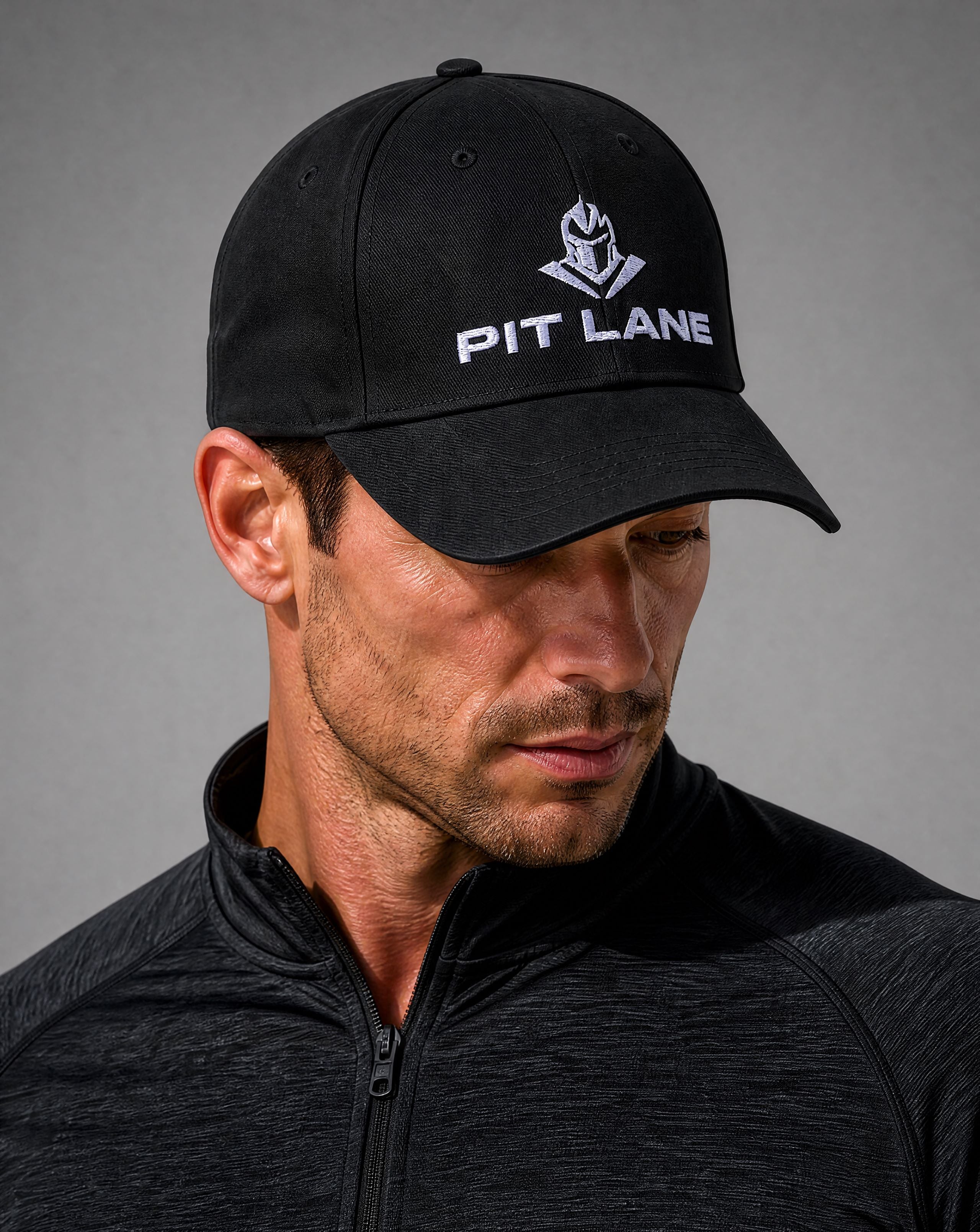 A man wearing a black baseball cap with a white embroidered PIT LANE logo and shield emblem, paired with a dark gray athletic zip-up jacket against a neutral gray background.