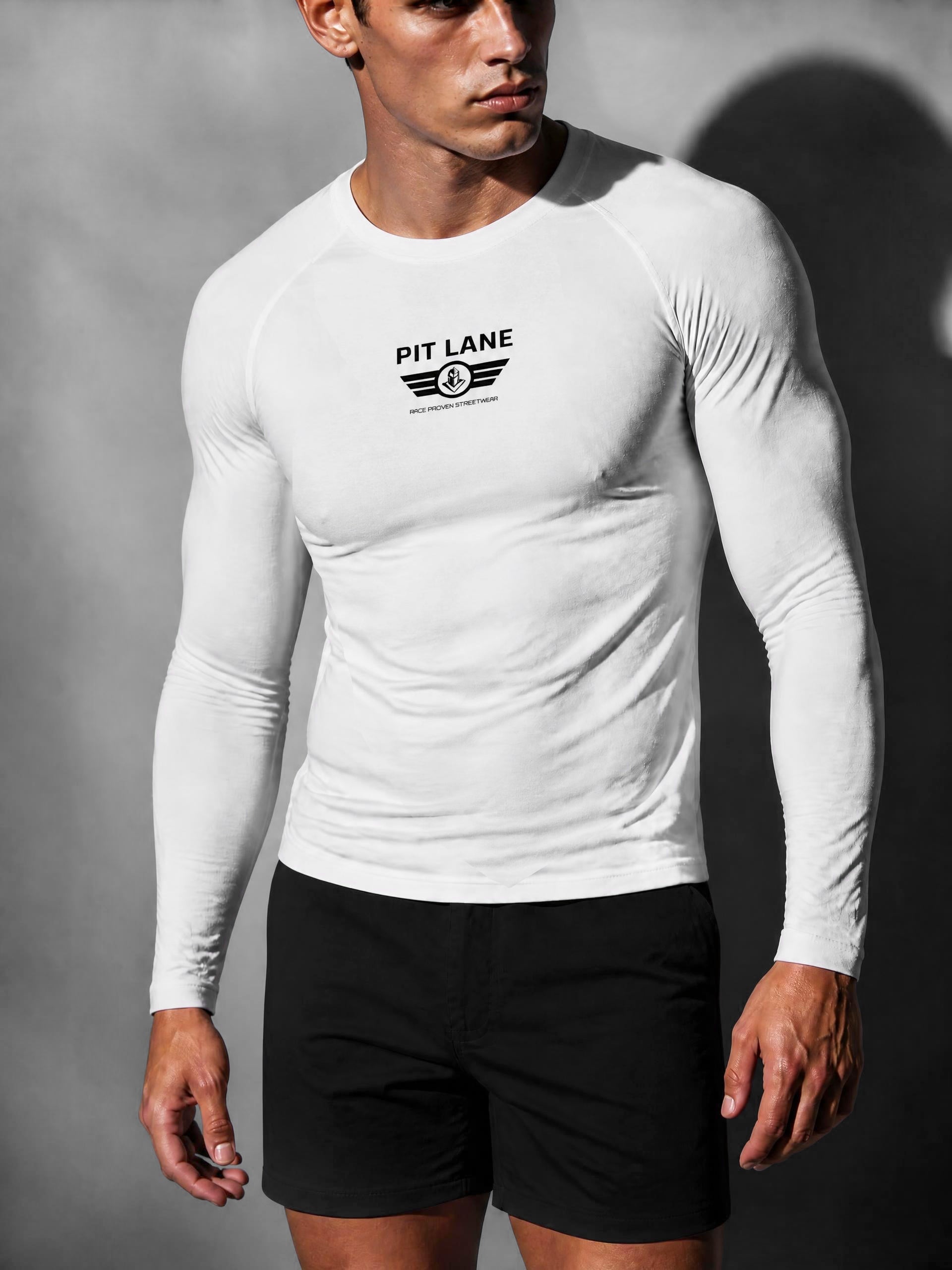 A man wearing a fitted white long-sleeve athletic shirt with a black 'PIT LANE' chest logo, paired with black shorts, photographed against a dark gray studio background.