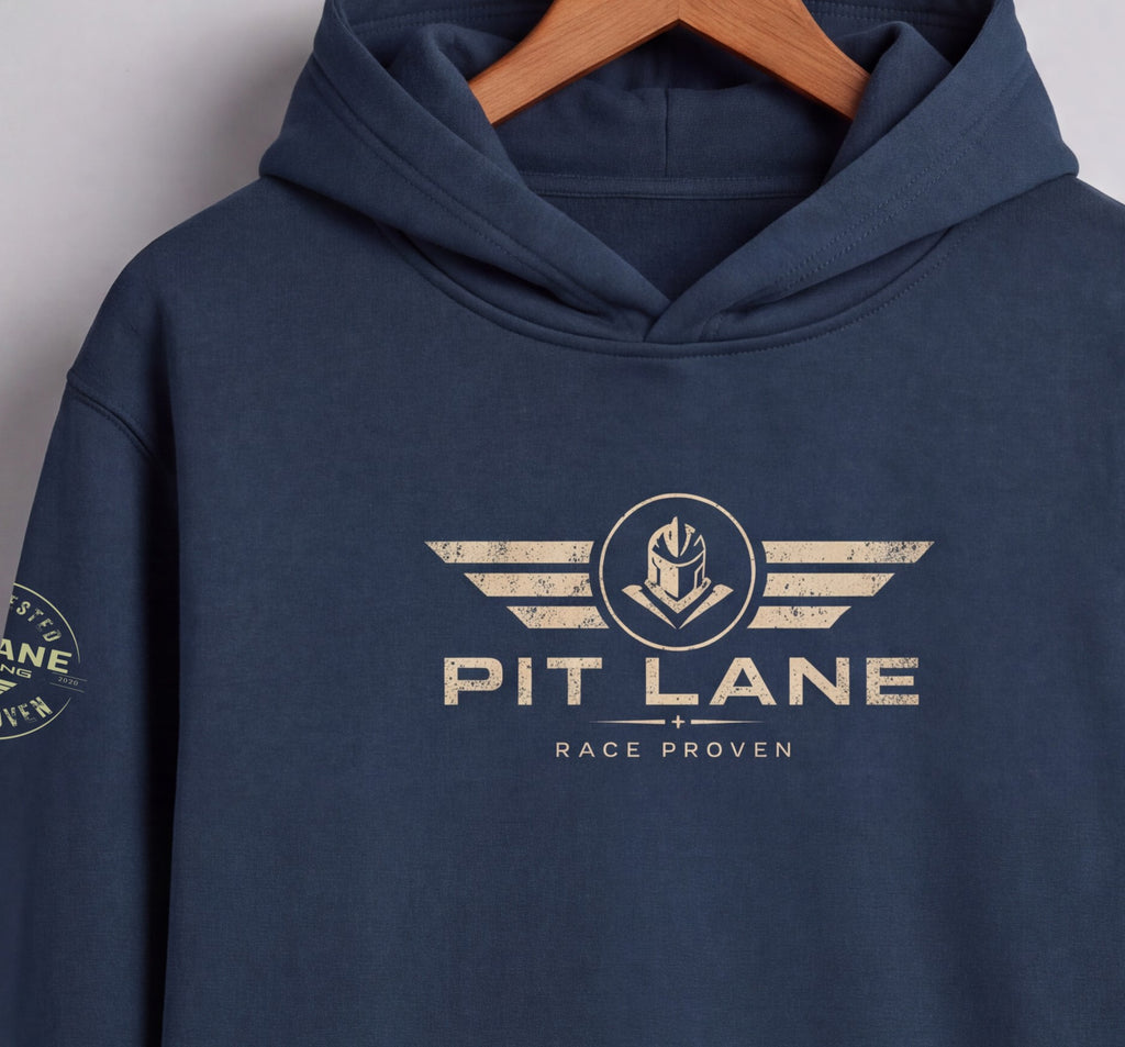 A navy blue pullover hoodie hanging on a wooden hanger against a light gray wall. The hoodie features a distressed beige graphic on the chest with the text 'PIT LANE' and 'RACE PROVEN' beneath it, and a matching circular graphic is visible on the left sleeve.