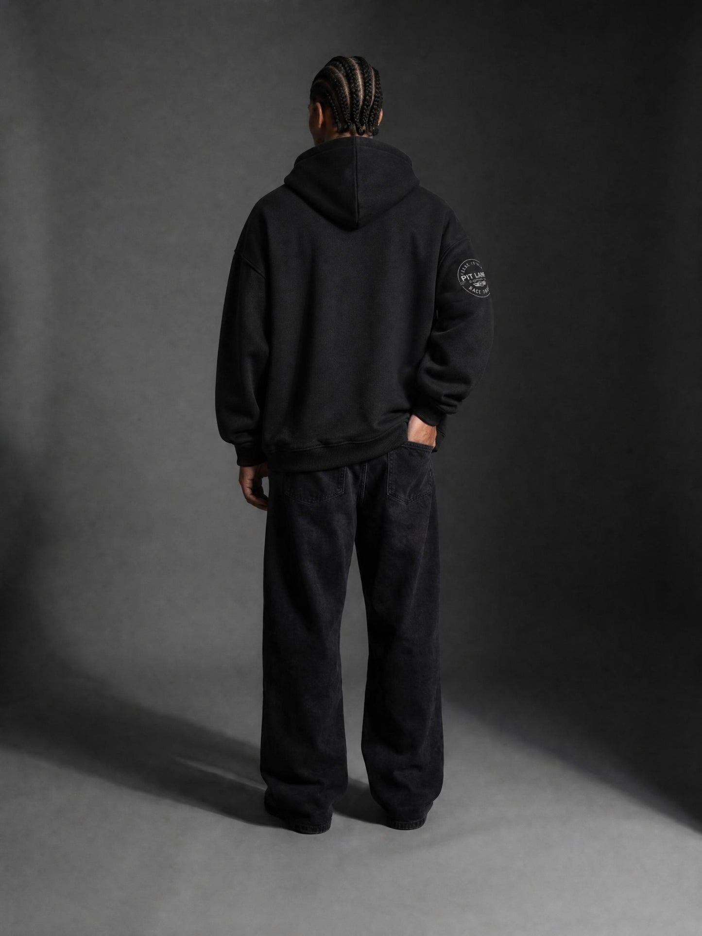 A man wearing a PIT LANE CLOTHING black pullover hoodie, paired with black sweatpants and black sneakers, standing against a dark gray studio background.