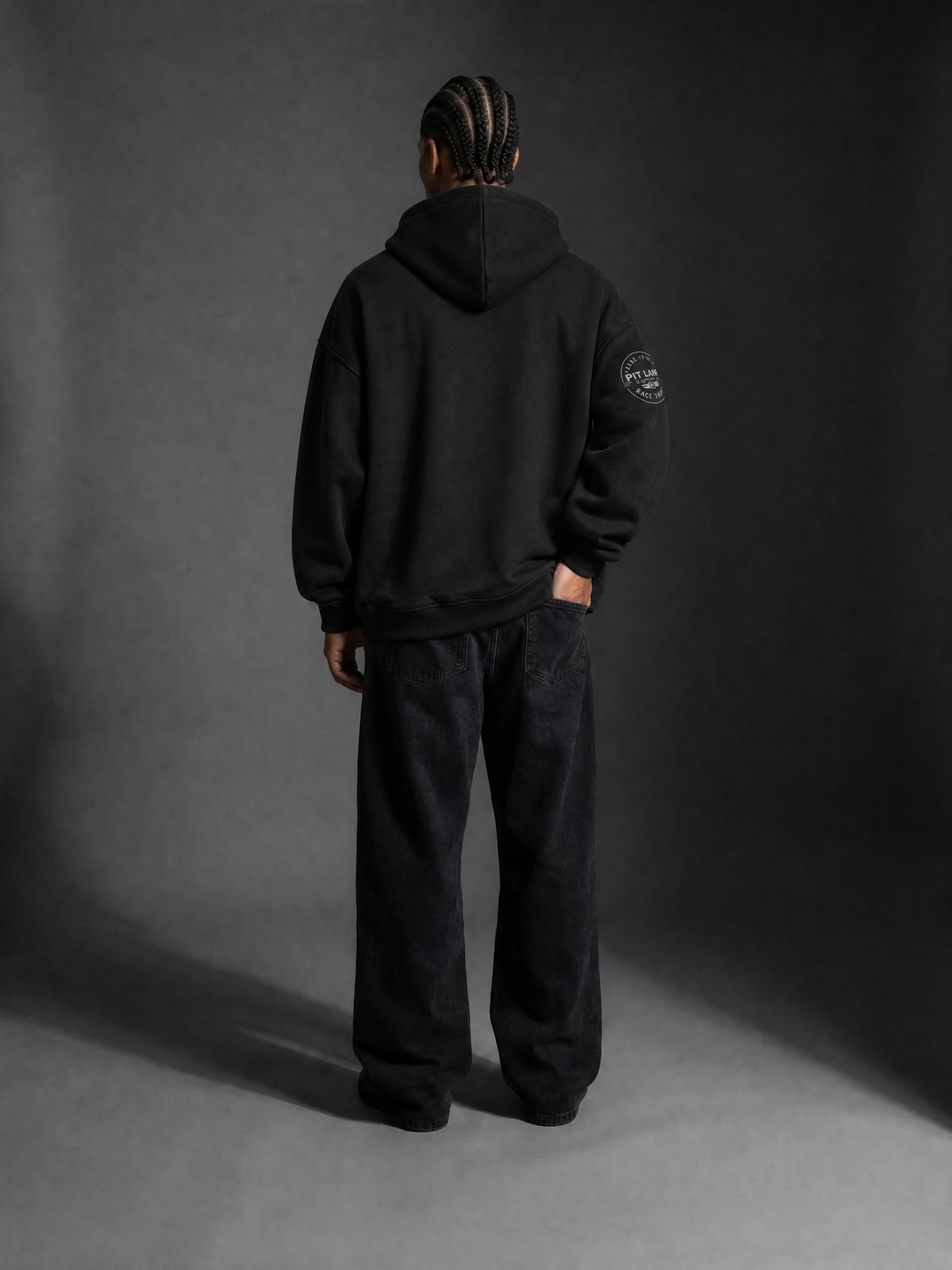 A man wearing a PIT LANE CLOTHING black pullover hoodie, paired with black sweatpants and black sneakers, standing against a dark gray studio background.