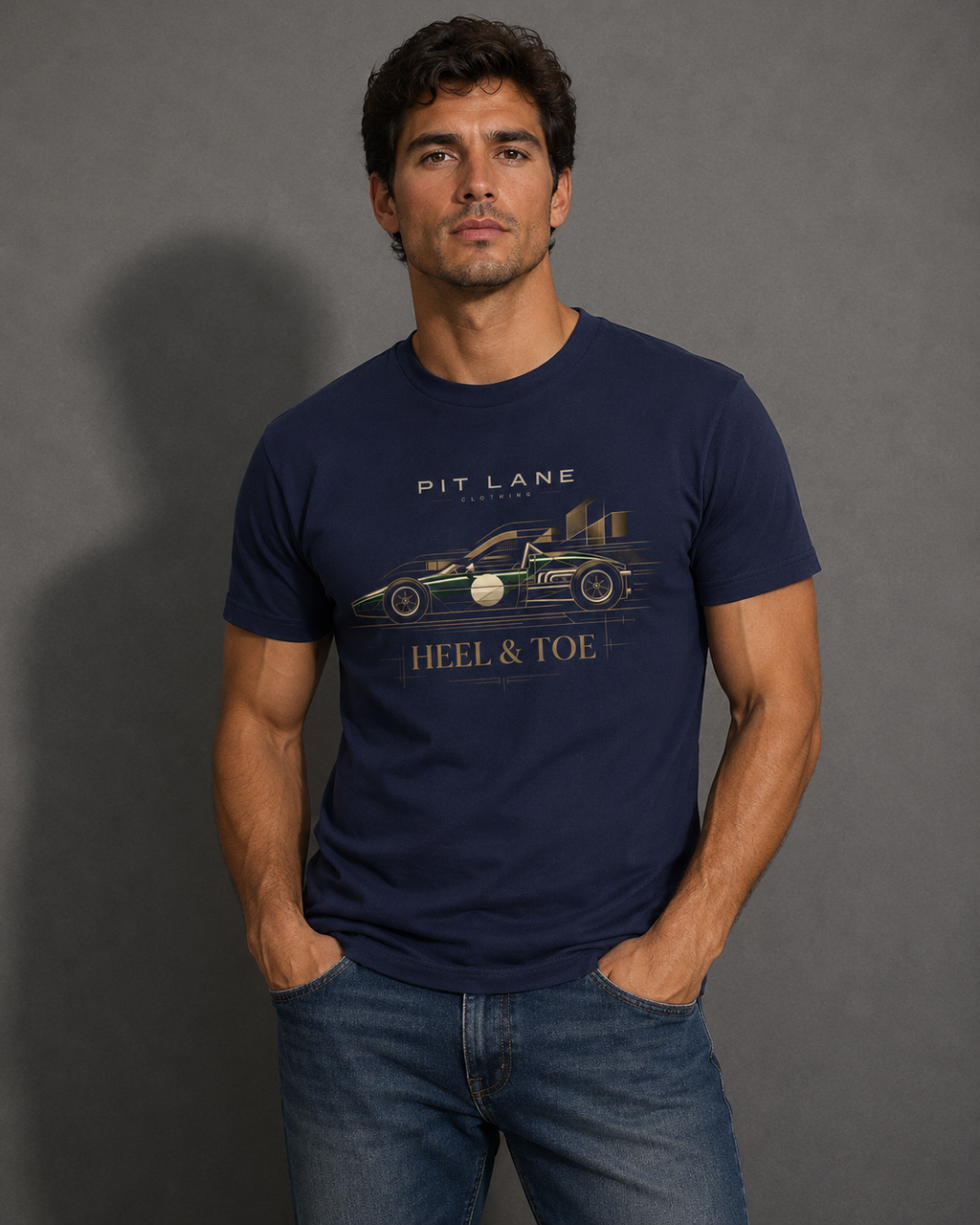 A man wearing a navy blue graphic t-shirt with a vintage race car and 'PIT LANE CLOTHING HEEL & TOE' text, paired with blue jeans, standing against a dark gray studio background.