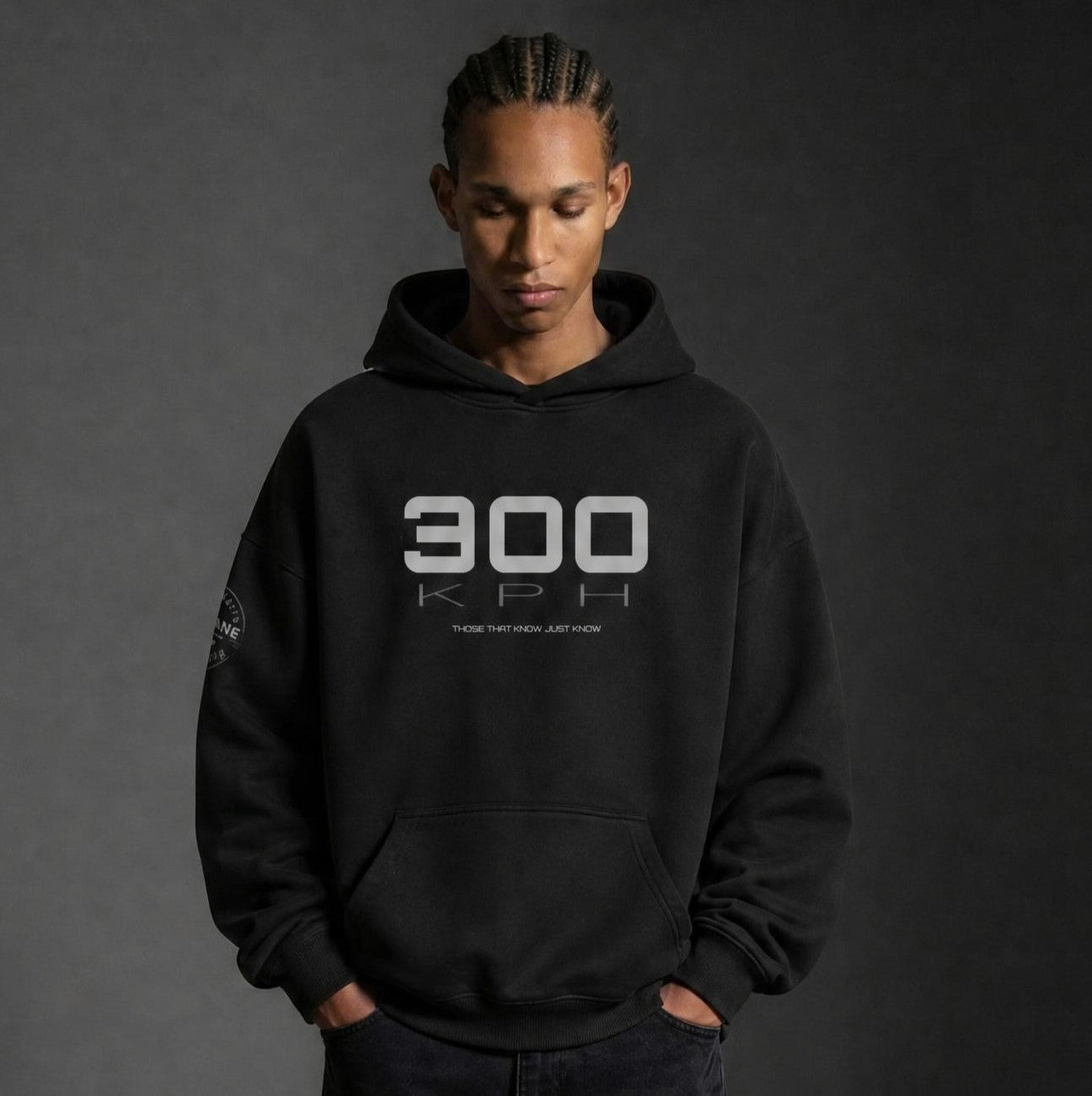 A man wearing a PIT LANE CLOTHING black pullover hoodie with a white '300 KPH' graphic on the chest, paired with black sweatpants and black sneakers, standing against a dark gray studio background.