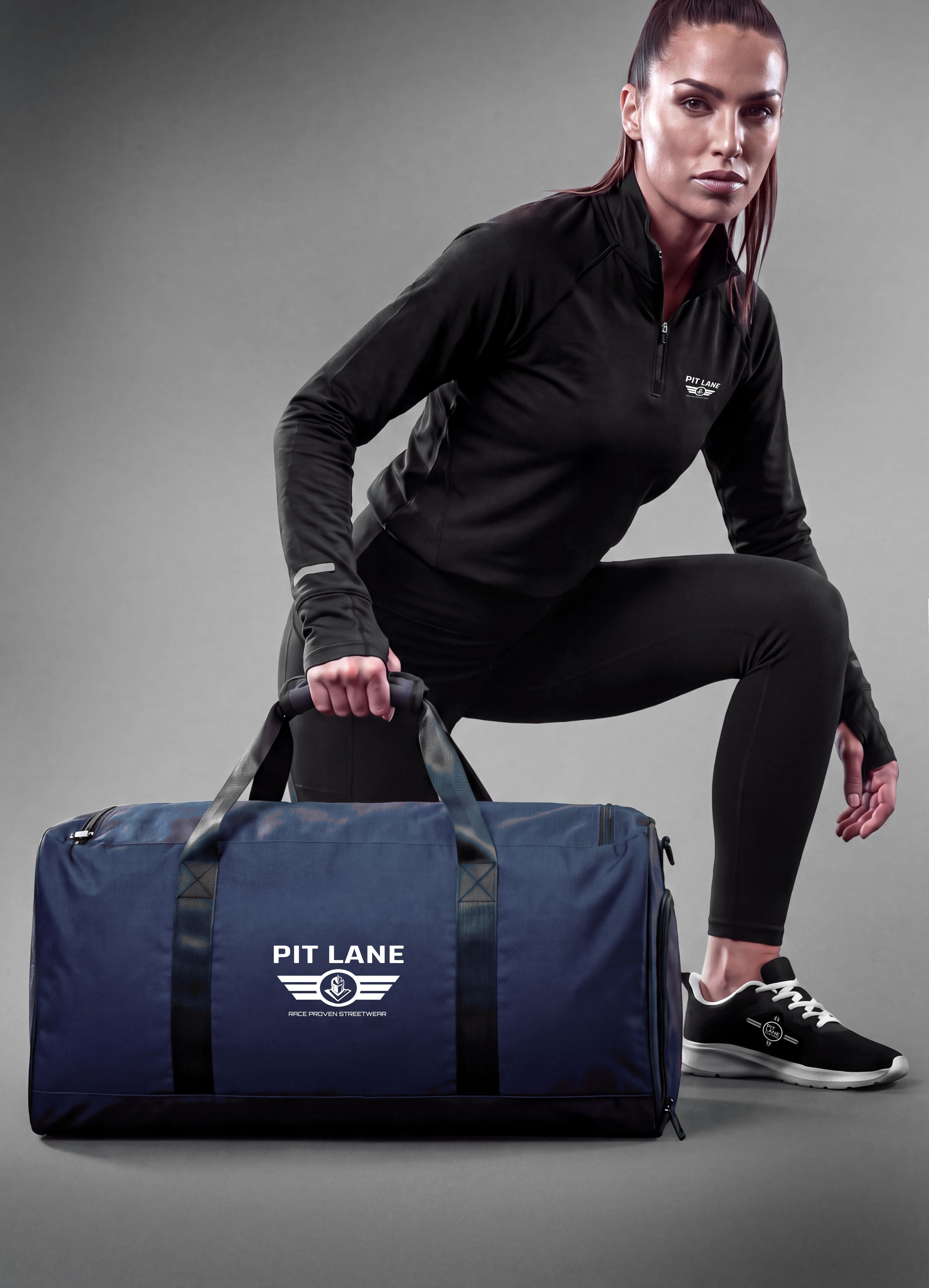 A woman in a black athletic outfit crouches beside a navy blue duffel bag with black handles and a white 'PIT LANE' logo on a gray studio background.