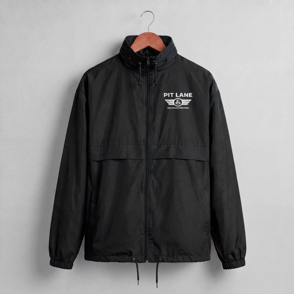 A black hooded windbreaker jacket hanging on a wooden hanger against a light gray wall. The jacket features a white 'PIT LANE' winged logo embroidered on the left chest.