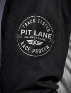 Close-up of a black garment with a circular white graphic print reading 'PIT LANE CLOTHING', 'TRACK TESTED', and 'RACE PROVEN'.