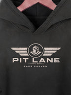 A close-up of a black hooded sweatshirt hanging on a pink hanger, featuring a distressed white graphic on the chest with wings, a helmet emblem, and the text 'PIT LANE' and 'RACE PROVEN'.